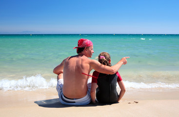 man and daughter sitting on sunny deserted beach