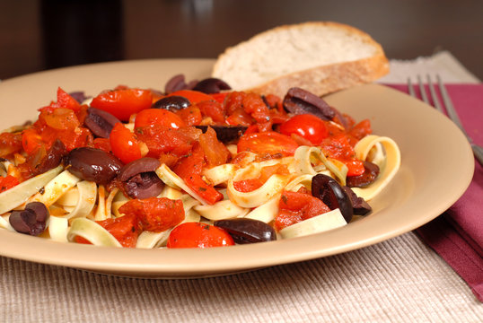 closeup of a plate of pasta puttanesca with wine and bread