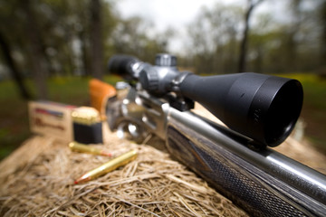 rifle with a rifle scope on hay with ammunition and trees behind. 