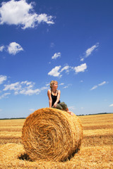 Naklejka na ściany i meble Woman sitting on a hay bale in stubble field under a bright sky