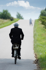 Old person in suit and hat riding a vintage bicycle.. - 3829404