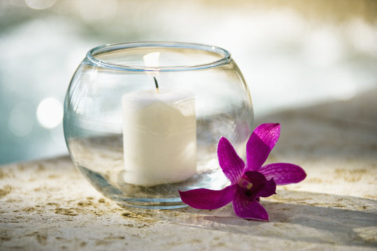 Lit candle in glass bowl and purple orchid beside pool.