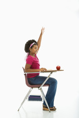 Side view of girl sitting in school desk raising hand.