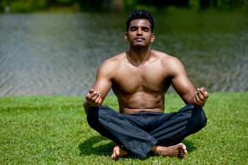 A well-built Asian man practising yoga outside by water