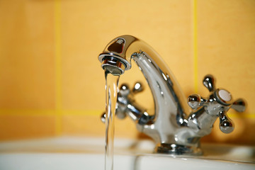 Working water tap. Flowing water. Selective focus, shallow depth of field. Blurred yellow tiled...