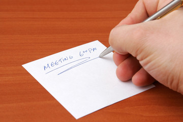 Hand holding a pen writing "Meeting a 6 PM"