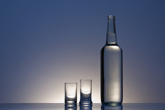 Clear Bottle With Two Vodka Glasses Against Blue Background.