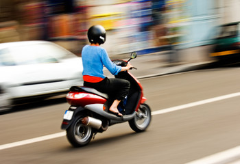 Panning shot of a young girl riding a scooter in a European city - obrazy, fototapety, plakaty
