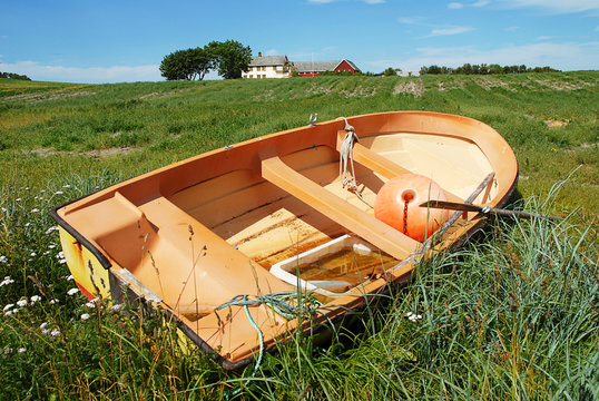 Boat towed ashore on green grass. In the background a farm on the horizon line.