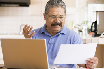 Man in kitchen with laptop and paperwork frustrated