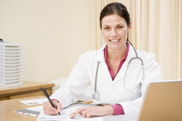 Doctor with laptop writing in doctor's office smiling