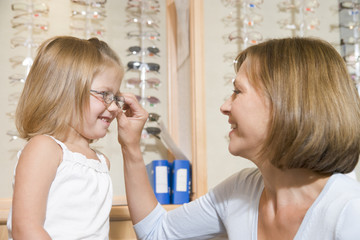 Woman trying eyeglasses on young girl at optometrists smiling