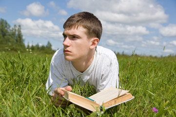 A young man reads a book, lying on a grass.