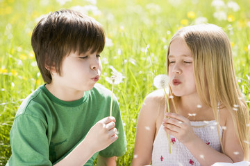 Two young children sitting outdoors blowing dandelion heads smil