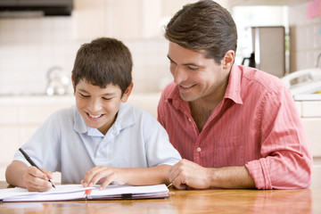 Man helping young boy in kitchen doing homework and smiling