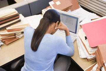 Businesswoman in cubicle with laptop and stacks of files