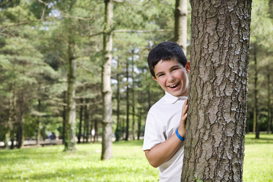 Portrait of young happy boy behind tree at park.