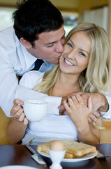 A young man kisses his wife whilst she is sat having breakfast