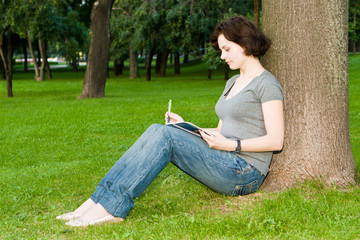 Girl sits under a tree in park and writes to notebook