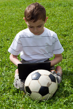 The boy with notebook and a football sits on a grass.