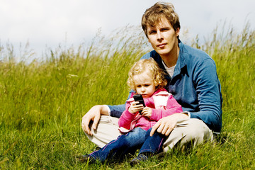 Father and child exploring mobile phone sitting in the grass.