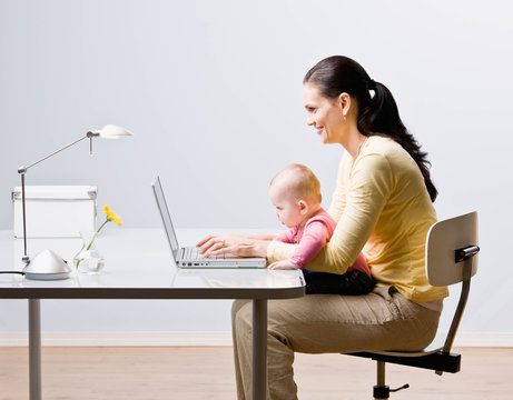 Mother holding baby while typing on laptop at desk