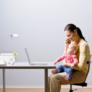 Working Mother Holding Baby While Talking On Cell Phone At Desk