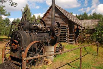 Old steam machine staying in a backyard, in the back old cottage - obrazy, fototapety, plakaty