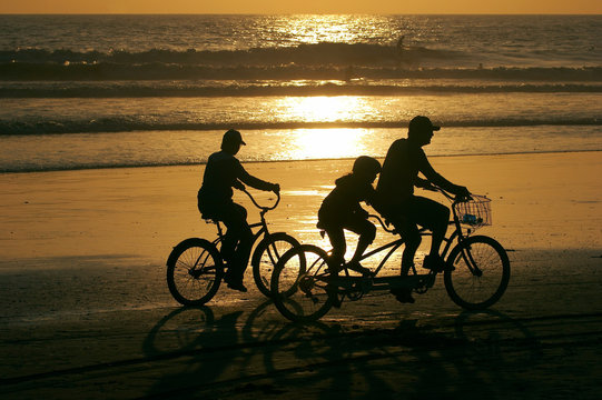 Family Riding Bikes Along The Beach At Sunset.