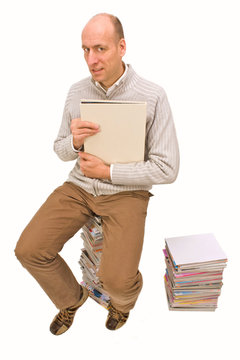 Mann sitzt auf einem Zeitschriftenstapel und schaut ein Buch an