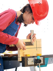 Eine junge Frau mit rotem Schutzhelm misst ein Stück Holz, das an einem Arbeitstisch befestigt...