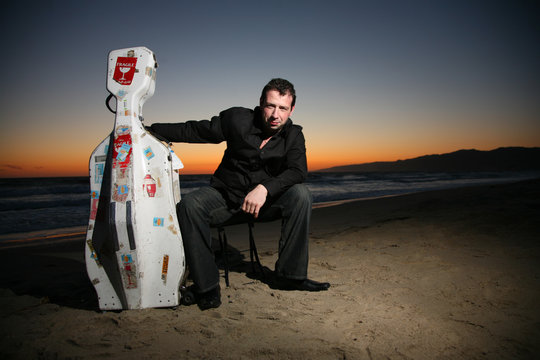 Musician With Travel Cello Or Guitar Case On Beach At Sunset