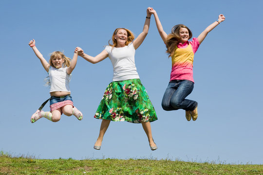 Happy Mother And Two Daughter Jumping On Green Meadow