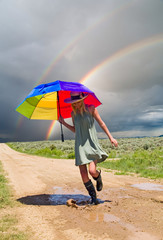 Girl with a rainbow umbrella splashing water in a puddle