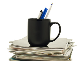 Pens in a coffee mug atop stack of magazines