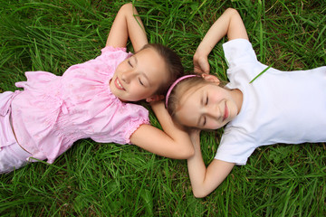 Two girls lie on grass with closed eyes,  head to head