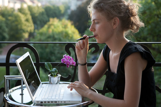 junge Frau schreibt auf einem Laptop auf dem Balkon, überlegt
