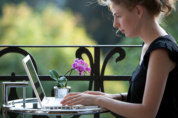 junge blonde Frau schreibt auf einem Laptop Computer auf Balkon - 16345692
