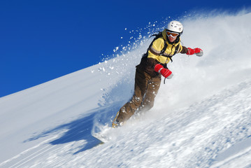 Young lady snowboarder in brown-yellow clothes riding down the slope and splashing snow