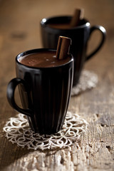 hot chocolate in black mugs with cinnamon stick, on old wooden t