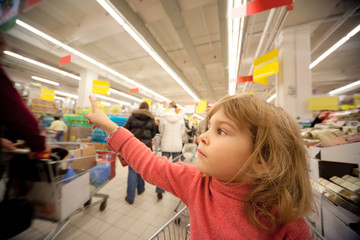 Small girl sit in shoppingcart in supermarket and show by finger