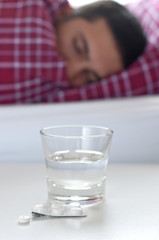 Tabletten und ein Glas Wasser auf Nachttisch vor Person im Bett