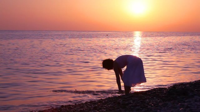 woman sloshing water in sea, sunset sky in background
