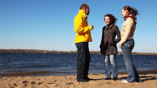 man and two woman stands on coastline of river talk and smile