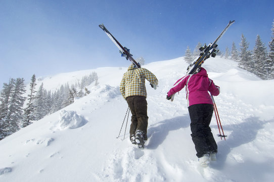 teenage couple (16-17) holding skis hiking up slope back view.