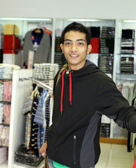 A handsome young Asian man wearing a casual hoodie posing inside a clothing store. He looks stylish and modern, browsing or preparing to shop for clothes. The setting is trendy and urban, suitable for