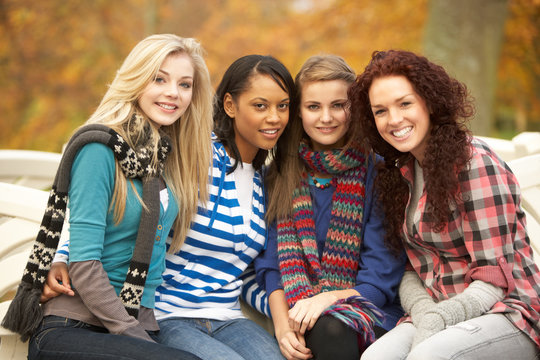 Group Of Four Teenage Girls Sitting On Bench In Autumn Park