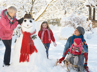 Young Girl With Grandmother And Mother Building Snowman In Garde
