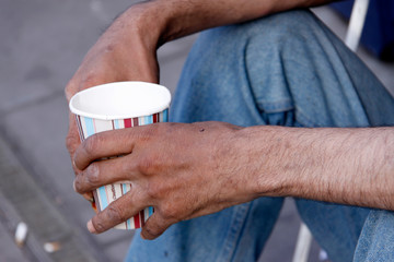 Close-up of anonymous beggar man holding out a paper cup while sat on the pavement 