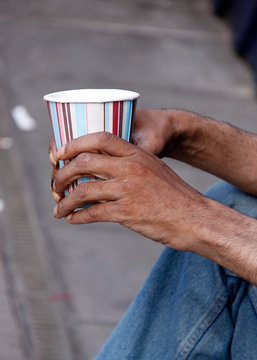 Anonymous beggar mans frail hands holding a cup out while sitting on pavement in the street 